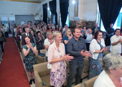 Audience applauding at an event in a historic hall with weathered walls, dark curtains, and rows of beige chairs.
