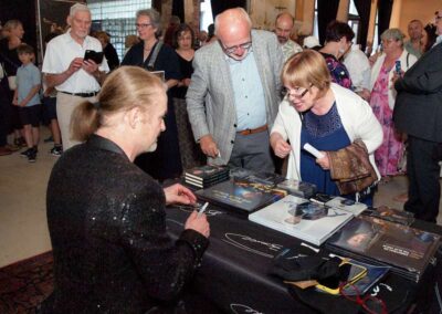 Musician in sequined jacket signing CDs and books for fans at an autograph event in a rustic venue