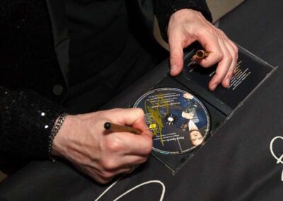 Person signing a CD with gold marker at autograph event, wearing black sequined jacket and beaded bracelet.