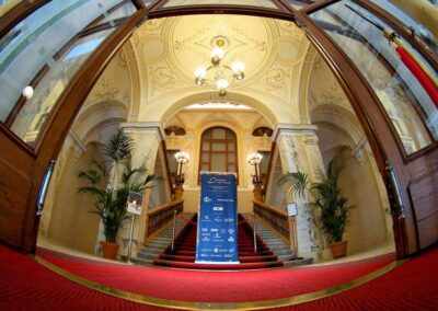 Ornate historic building lobby with grand staircase, red carpet, chandelier, and blue event banner display.