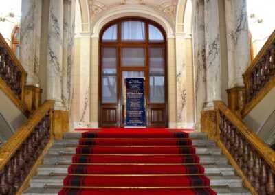 Ornate palace staircase with red carpet runner leading to grand doors, marble columns, and gilded arched ceiling.