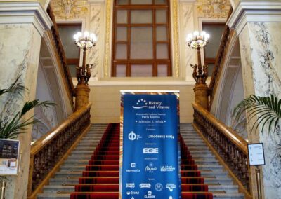 Ornate historic staircase with red carpet and blue Hvězdy nad Vltavou music festival banner in grand marble hall.