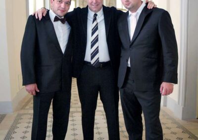 Three men in formal black suits posing together in an elegant hallway with arched windows and tiled floor.