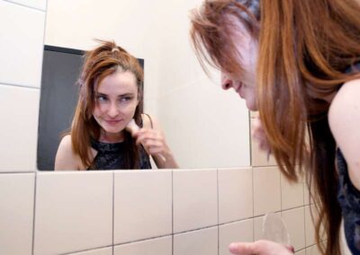 Woman with long auburn hair examining her skin in a tiled bathroom mirror, holding a contact lens case.