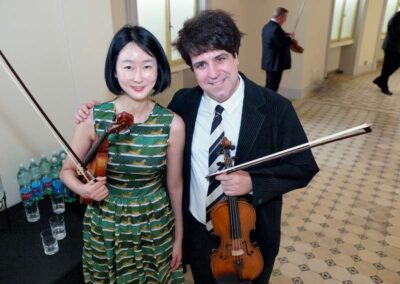 Two smiling violinists holding their instruments and bows pose together at an indoor event or concert venue.
