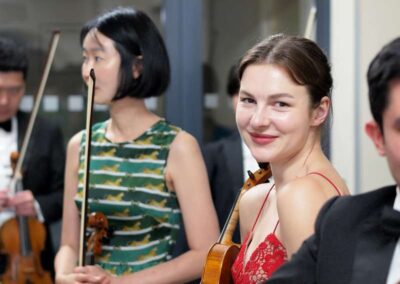 Smiling female violinist in red dress holding violin among orchestra musicians backstage before performance