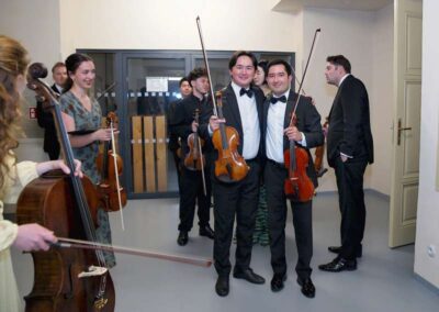 Young classical musicians in formal attire holding violins and cello, smiling together backstage before a concert performance.
