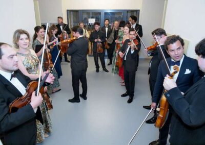 Chamber orchestra musicians in formal attire playing violins and holding instruments in a bright hallway before a performance.