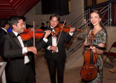 Two men in tuxedos playing violins beside a smiling woman in a floral dress holding a viola at a formal event.