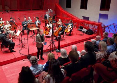 Chamber orchestra performing on red stage as speaker addresses seated audience in intimate concert hall venue