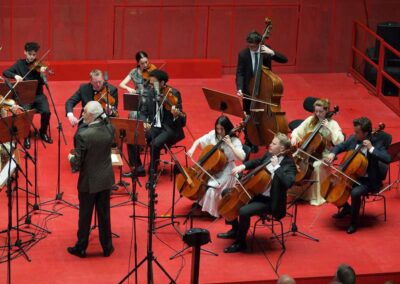 Chamber orchestra performing on red stage with conductor, cellists, violinists, and double bass player during live concert.