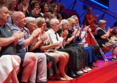 Audience members clapping enthusiastically while seated in a red-tiered auditorium at a live event.