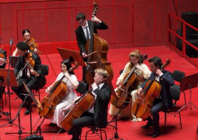Young orchestra musicians performing on stage with cellos, violins, and double bass against a striking red backdrop.
