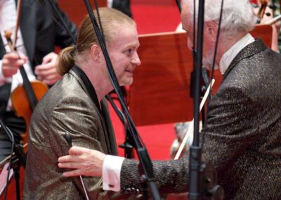 Two musicians in shimmering formal jackets greeting each other warmly at an orchestra concert stage with microphones.