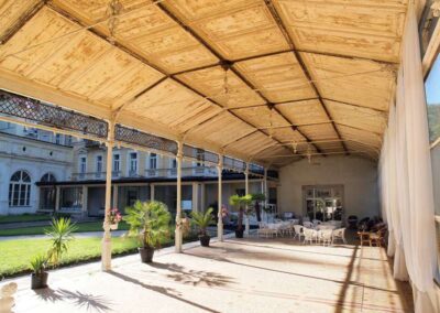Historic covered veranda with ornate wooden ceiling, iron columns, potted palms, and café seating beside a grand building.