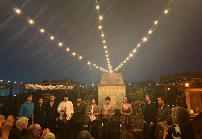 Chamber musicians with string instruments taking a bow under string lights at an outdoor evening concert.