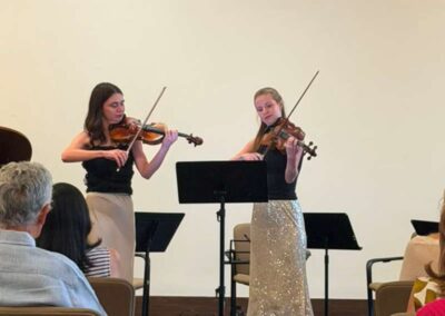 Two women playing violins during a recital in front of a seated audience with music stands.