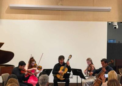 Chamber music ensemble with violins, cello, and classical guitar performing on stage before a seated audience