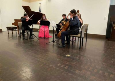 Chamber music ensemble performing with violins and cello, reading sheet music on stands in a recital hall.