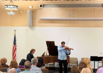 Violinist and pianist perform a classical music duet for a seated audience in an ornate wood-ceilinged recital hall.