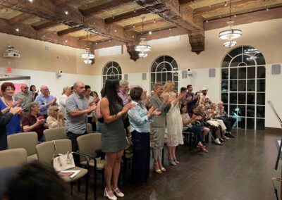 Audience standing and applauding at an indoor event in an elegant hall with ornate wood ceiling and chandeliers.