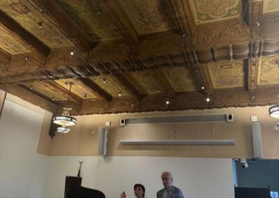 Two speakers presenting near a grand piano beneath an ornate carved ceiling at a Beverly Hills event venue.