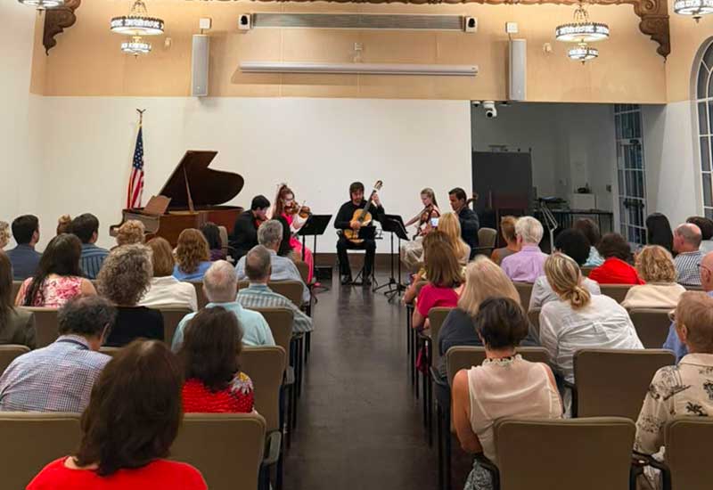 Classical guitarist performs with string ensemble before seated audience in an intimate concert hall recital.