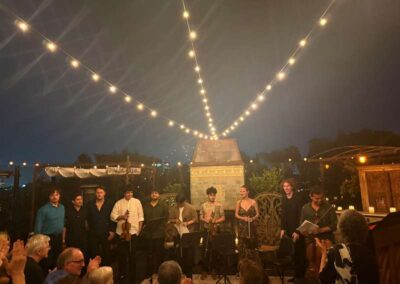 Musicians with instruments standing together on outdoor stage under string lights at evening concert as audience applauds