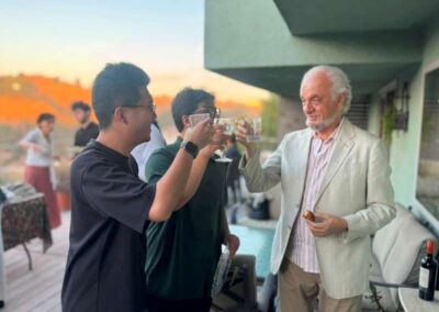 Three men toasting drinks at an outdoor sunset gathering on a balcony with a table of bottles and glasses.