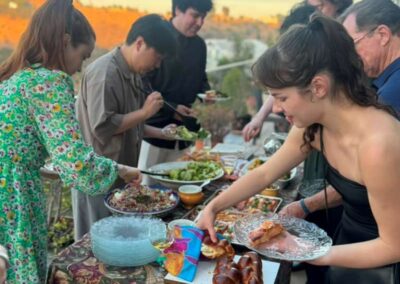 Guests serve themselves from a buffet table with challah bread and salads at an outdoor rooftop dinner party at sunset.