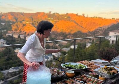 Woman setting up an outdoor buffet table with salads and entrées on a hillside patio at sunset.