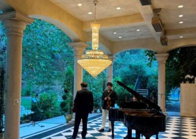 Musicians with violins and grand piano performing on checkered floor patio with chandelier and garden views.