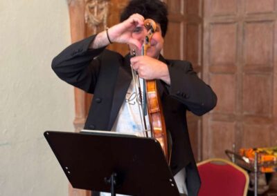 Violinist in black tuxedo tuning violin while standing behind a music stand in an ornate wood-paneled room.