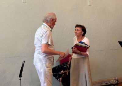 Two elderly musicians talking and exchanging sheet music at a rehearsal, with music stands and instrument case nearby.