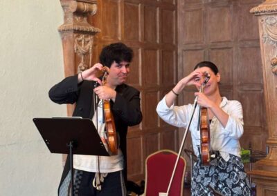 Two violinists tuning their instruments in an ornate wood-paneled room with music stands during a rehearsal.