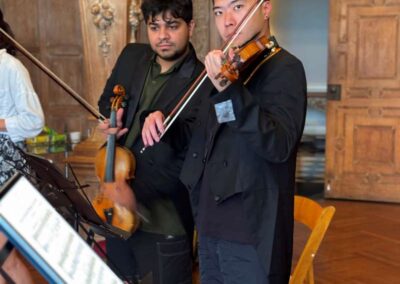 Two male violinists in black suits performing in an elegant wood-paneled room with ornate columns and sheet music stands.