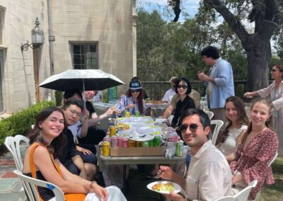 Group of friends enjoying an outdoor lunch gathering at a stone mansion, with drinks and food on a long table.