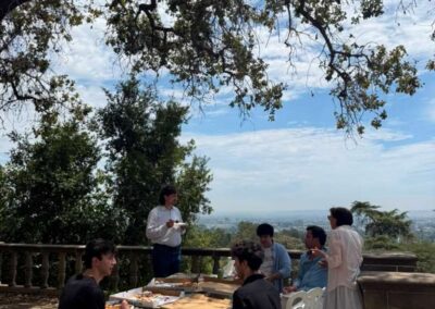 Group of friends enjoying pizza on a scenic hilltop patio overlooking a city under a large oak tree.
