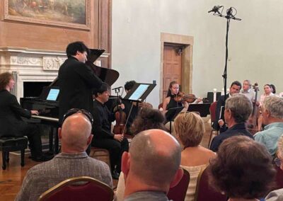 Chamber music ensemble performing with pianist and string players before an audience in an elegant historic hall.