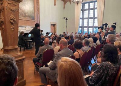 Classical music concert with pianist and string musicians performing for seated audience in elegant historic hall.