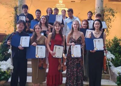 Music competition winners holding certificates and violins on garden steps decorated with white orchids and flowers.