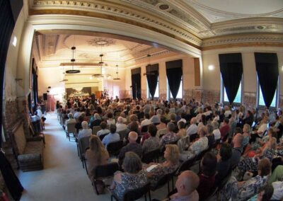Audience seated in ornate historic hall watching performance on stage with decorative ceiling and tall arched windows.