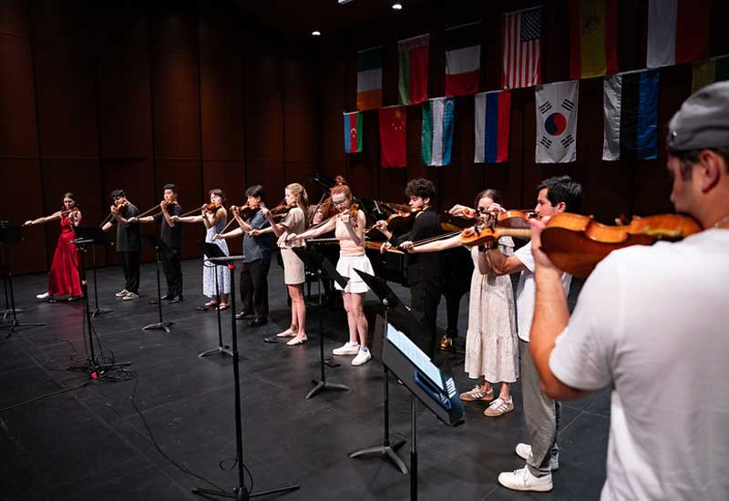 Violinists perform on stage with international flags displayed above during a music competition or masterclass.